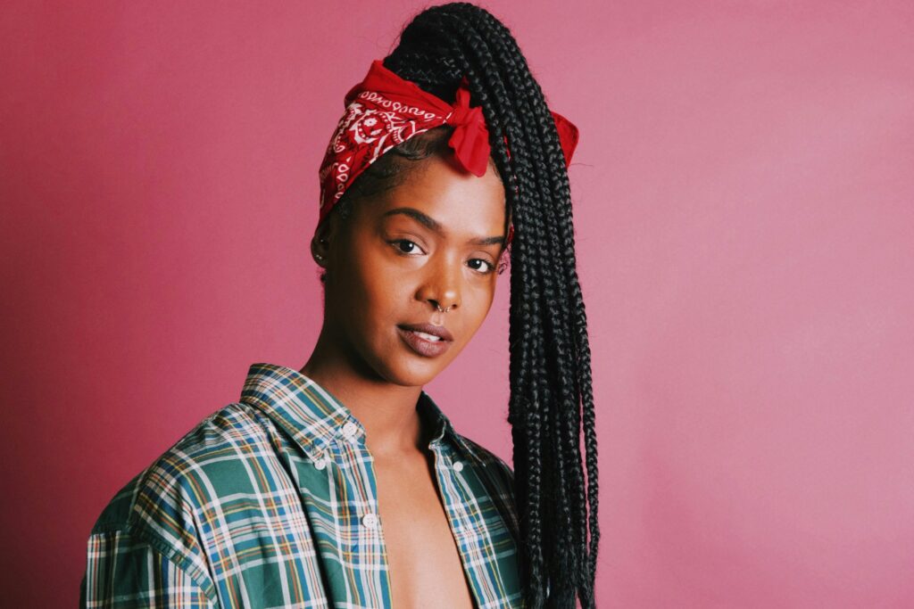 Portrait of a Black woman wearing long box braids styled in a high ponytail with a red bandana against a pink background.